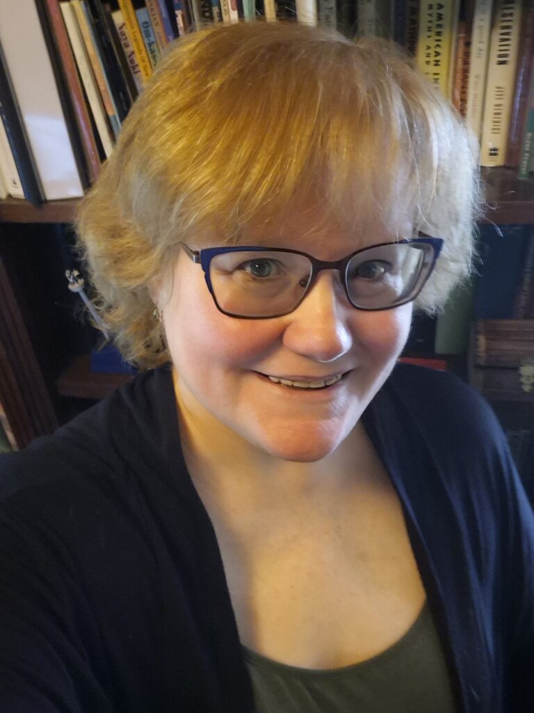 Image of Nancy C Walker. A pale woman with short, light red hair and blue eyes behind orange and blue rimmed glasses, wearing an army green shirt and a black  wrap in front of a bookcase.