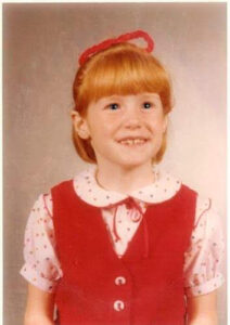 An image of a 6 year old girl with pale skin and bright read hair, where a pink shirt and red vest smiling at the camera.